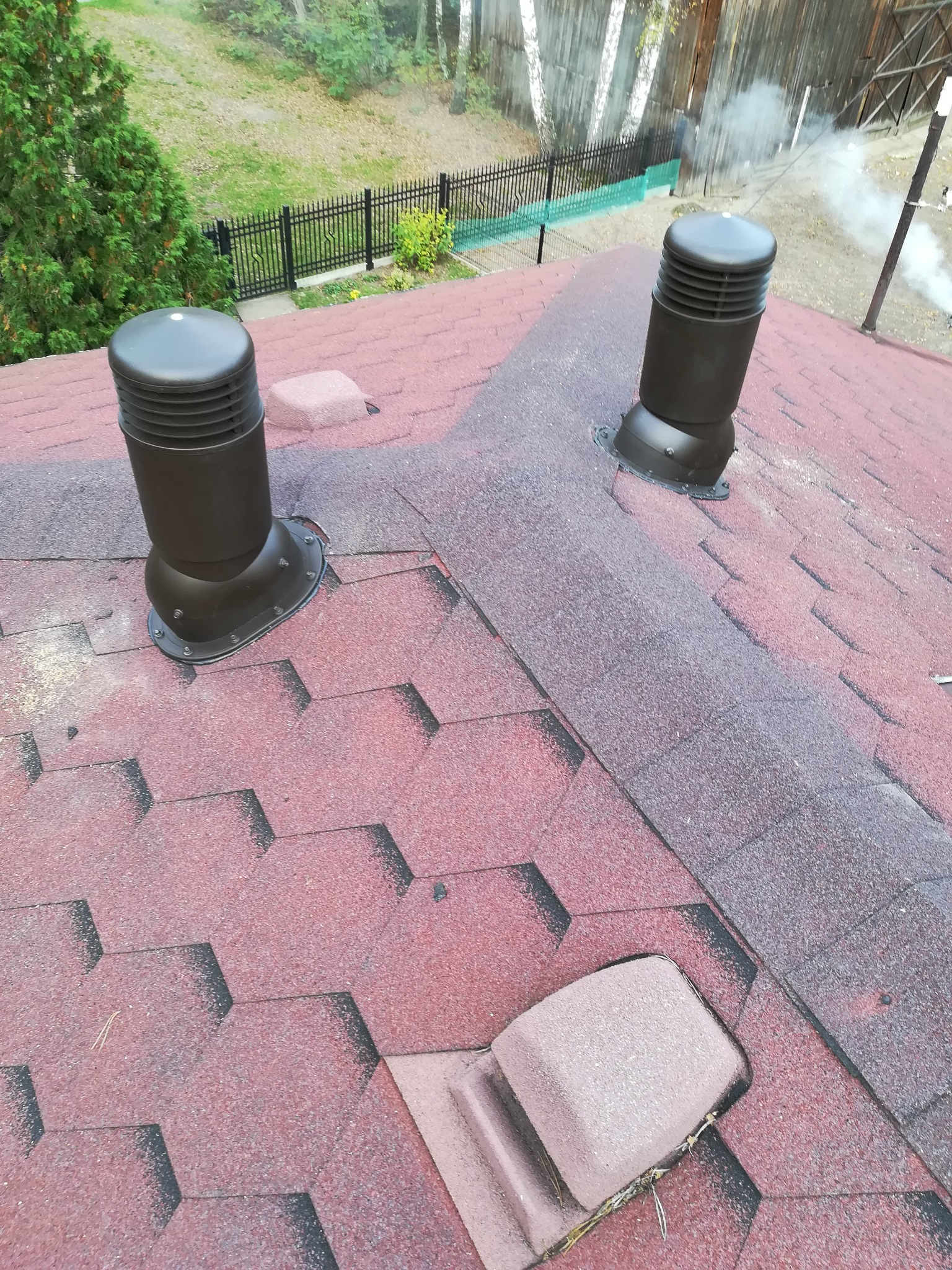 Widok z góry na dach pokryty czerwoną dachówką bitumiczną z dwoma brązowymi kominkami wentylacyjnymi, w tle drzewa i ogrodzenie.