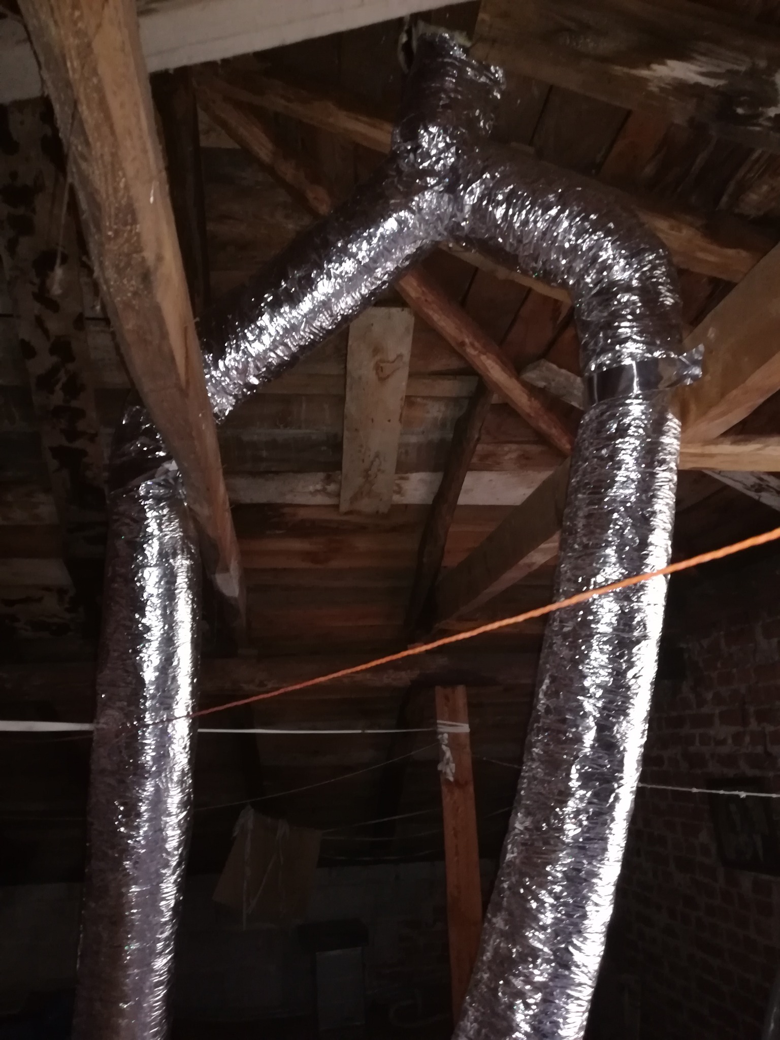 Srebrne, elastyczne rury wentylacyjne w przestrzeni poddasza, widoczne drewniane krokwie i ceglany mur.