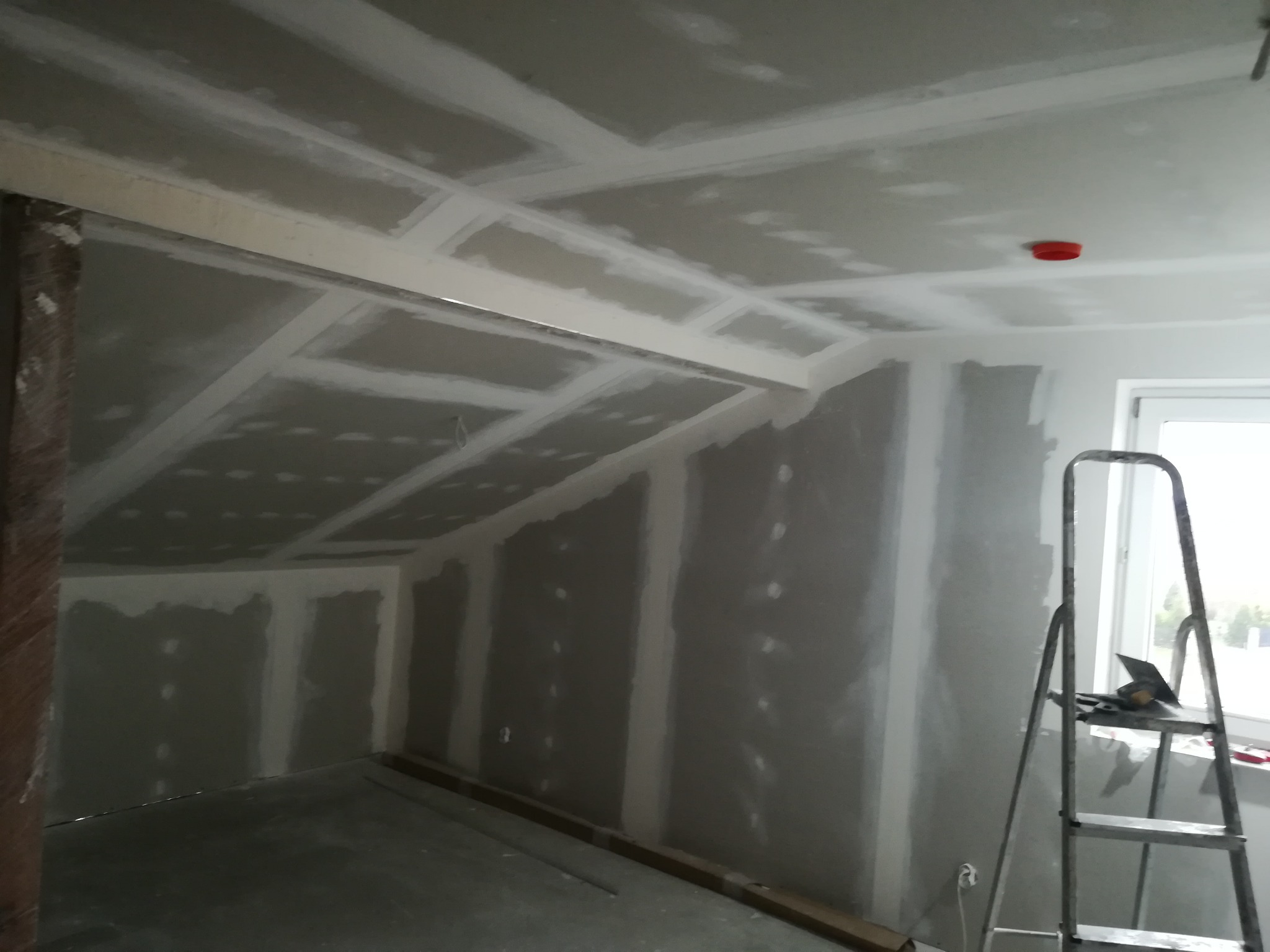 Poddasze w trakcie wykańczania, ściany i skośny sufit pokryte szarymi płytami, widoczne łączenia i szpachlowanie, drabina aluminiowa przy ścianie z narzędziami.