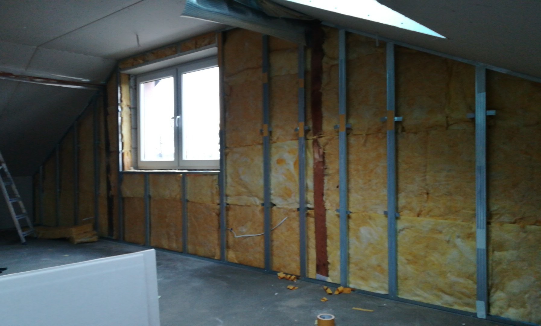 Montaż izolacji z wełny mineralnej w ścianie poddasza z oknem, widoczne profile metalowe i fragment podwieszanego sufitu