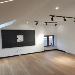 Jasne panele podłogowe w nowocześnie wykończonym pokoju na poddaszu z oknem, czarną listwą przypodłogową, czarnym panelem dekoracyjnym z wyciętym otworem na ścianie i reflektorami sufitowymi...