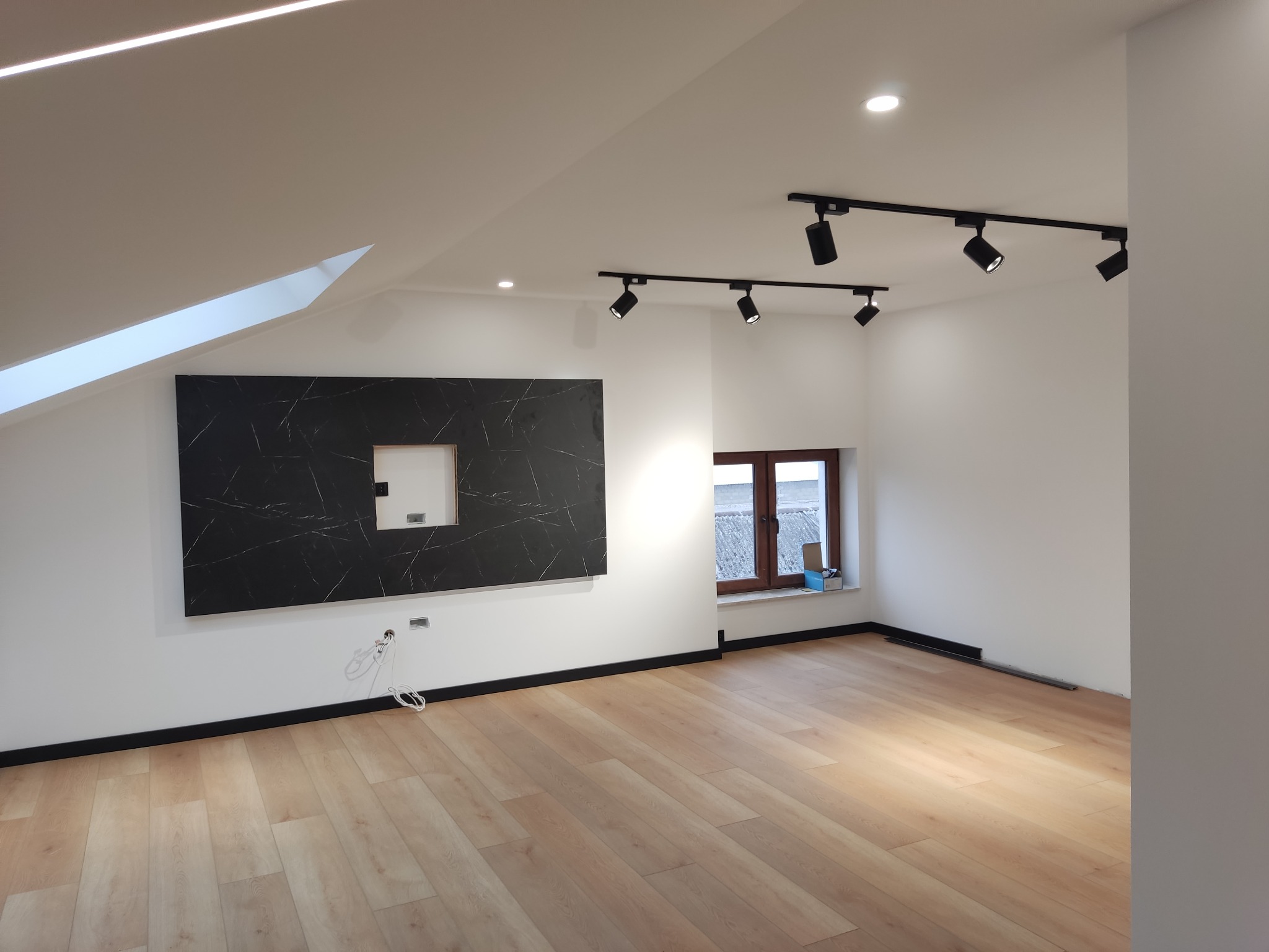 Jasne panele podłogowe w nowocześnie wykończonym pokoju na poddaszu z oknem, czarną listwą przypodłogową, czarnym panelem dekoracyjnym z wyciętym otworem na ścianie i reflektorami sufitowymi...