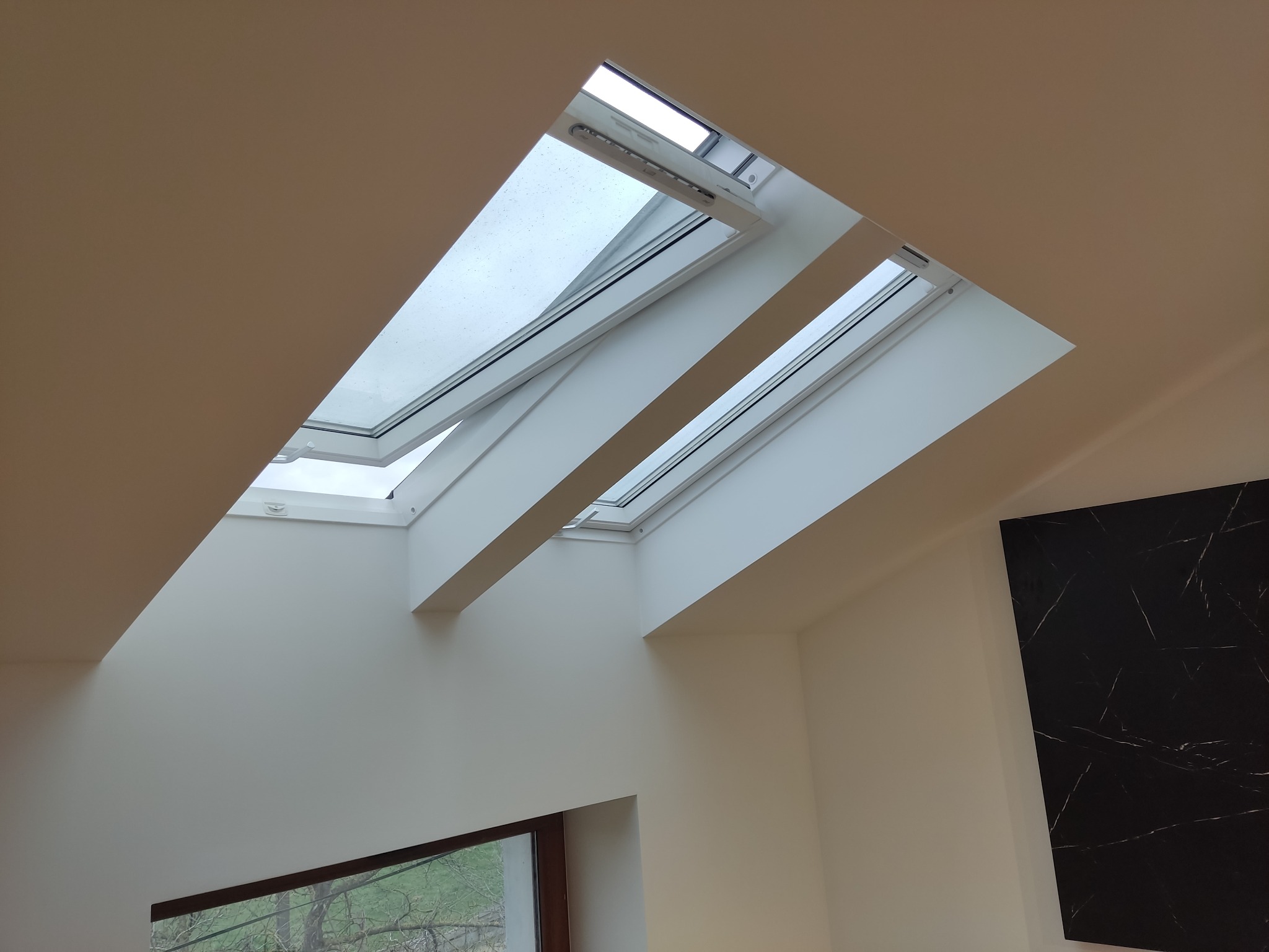 Wnętrze z trzema oknami dachowymi, częściowo otwartymi, widok na zewnątrz z kroplami deszczu na szybach, ściany w odcieniach bieli i beżu, dekoracyjny panel imitujący czarny marmur w rogu...