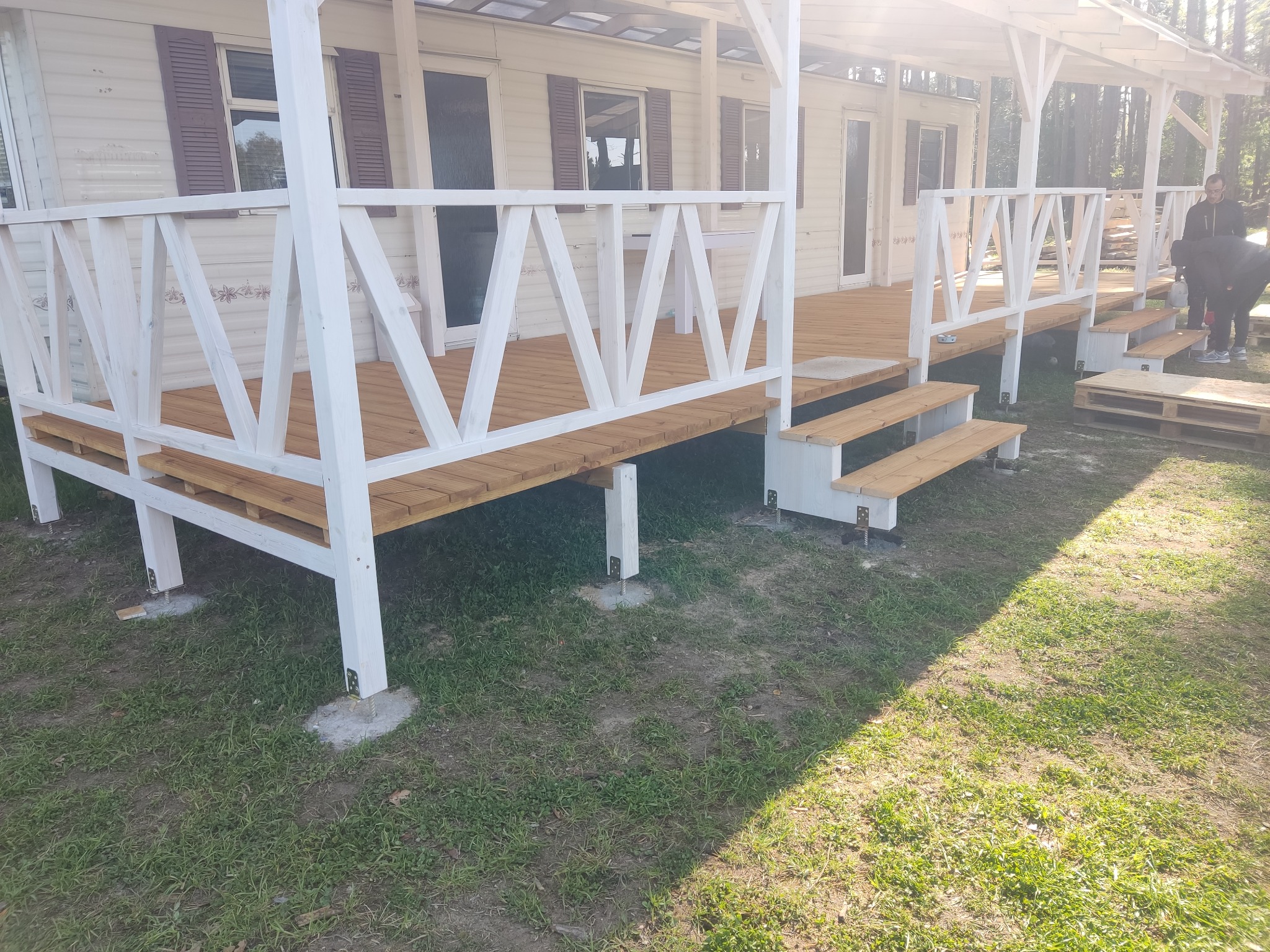 Zadaszony drewniany taras z białą balustradą przy domu mobilnym, widoczne betonowe podstawy pod konstrukcją, fragment lasu w tle.