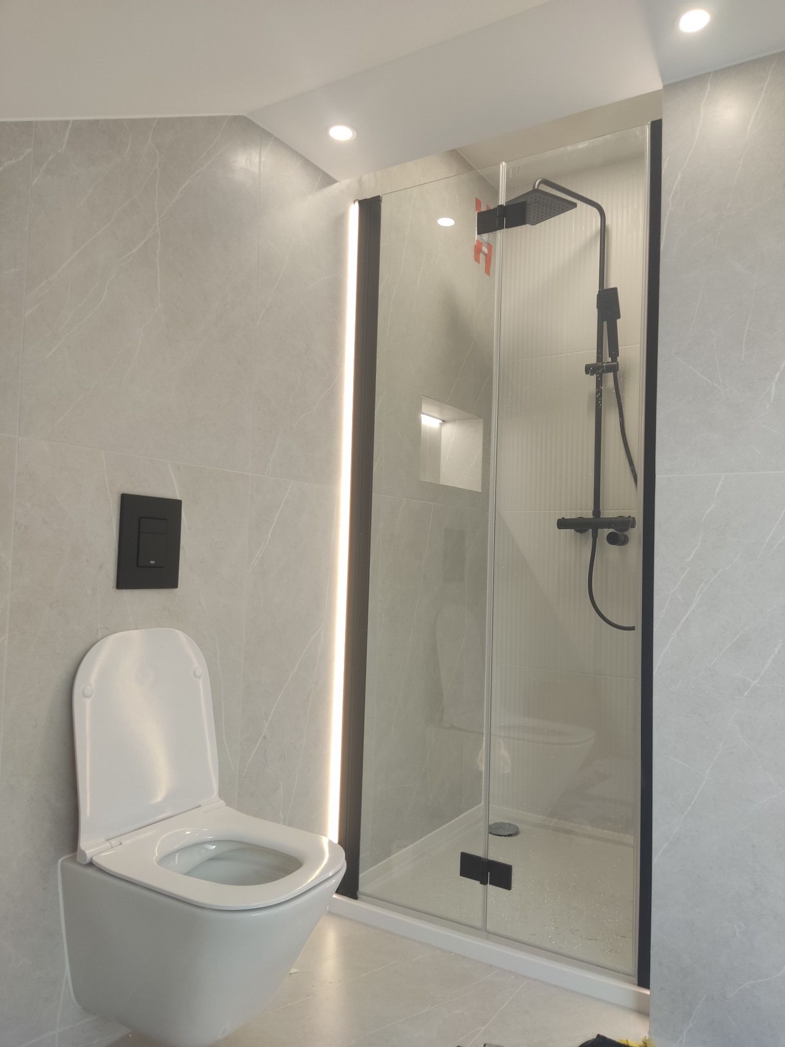 Nowoczesna łazienka z szarymi płytkami imitującymi marmur, wisząca toaleta z czarnym przyciskiem spłukującym i kabina prysznicowa typu walk-in z czarnymi profilami i deszczownicą.