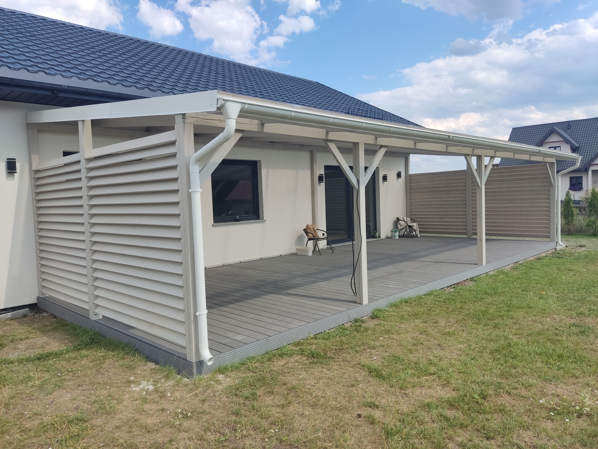 Zadaszony taras z szarych desek kompozytowych, częściowo osłonięty drewnianymi żaluzjami, z widocznym systemem rynnowym i elementami oświetlenia zewnętrznego.