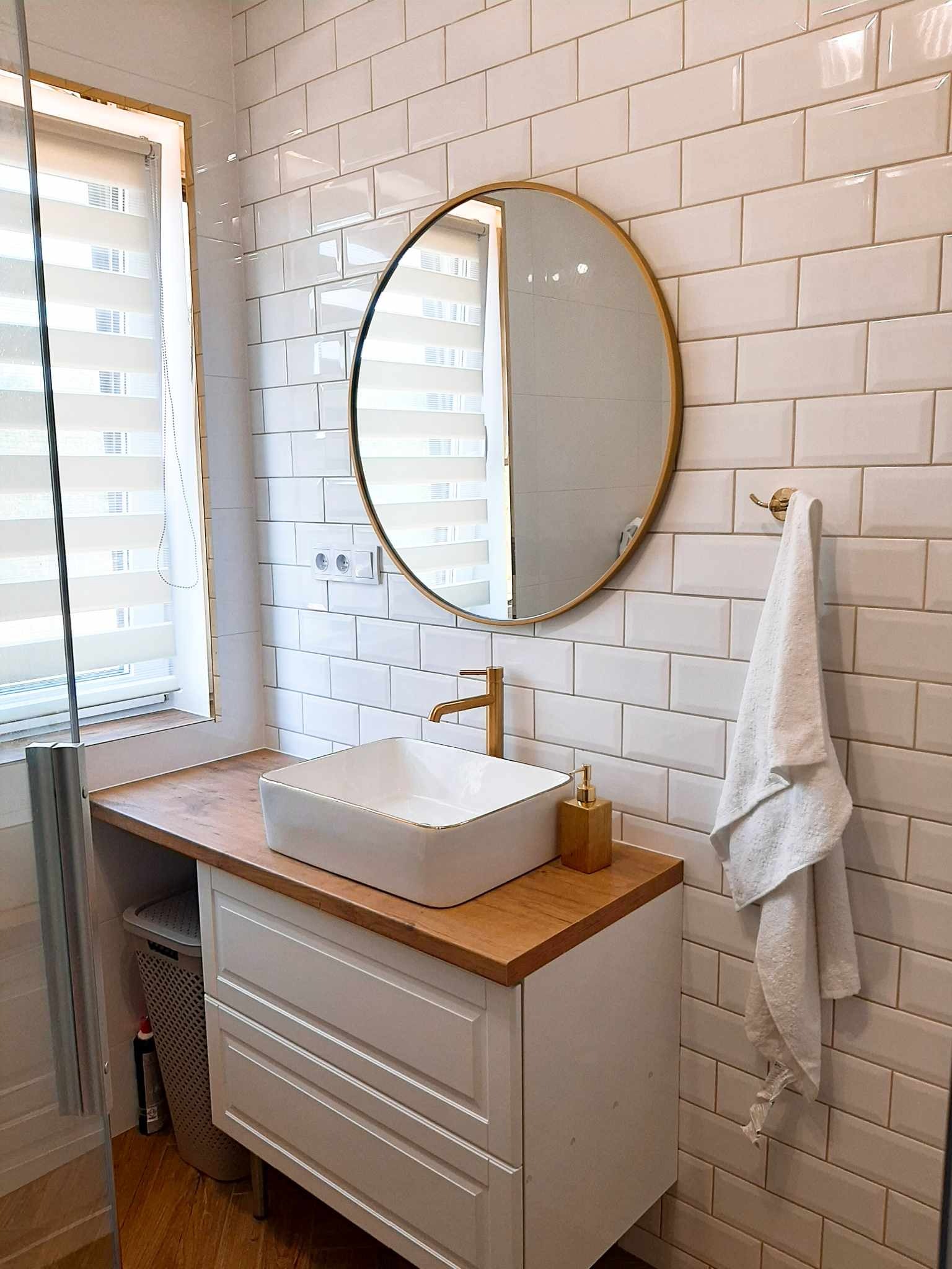 Biała, prostokątna umywalka z baterią w kolorze złotym, osadzona na drewnianym blacie z szafką w łazience z białymi, błyszczącymi kaflami typu metro i okrągłym lustrem w złotej ramie.