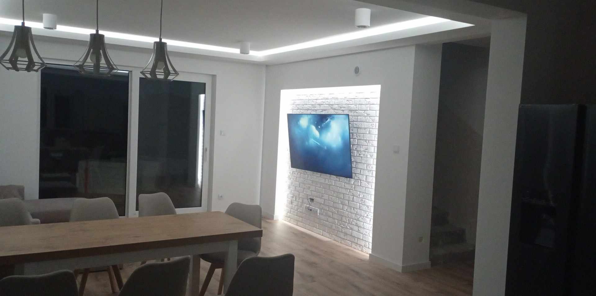 Jasne, nowoczesne wnętrze z drewnianym stołem, szarymi krzesłami i ścianą z białej cegły, na której wisi telewizor podświetlony od tyłu. Sufit z oświetleniem LED.