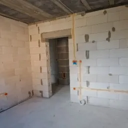 Surowe wnętrze w trakcie prac wykończeniowych, widoczne białe ściany z betonu komórkowego z wyfrezowanymi bruzdami pod przewody elektryczne i zamontowanymi puszkami elektrycznymi; sufit w stanie...