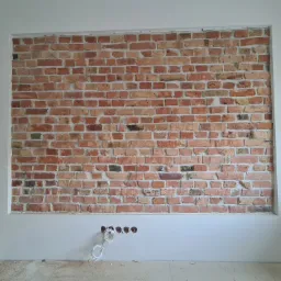 Fragment ściany z dekoracyjną cegłą, otoczony pomalowanym na biało fragmentem, w dolnej części widoczne otwory na kable.