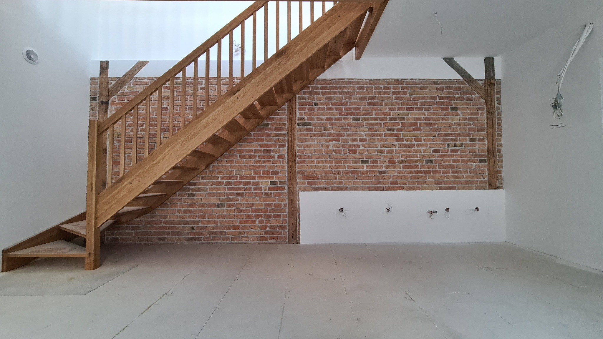 Surowe drewniane schody w nowym domu z ceglaną ścianą, widoczne instalacje elektryczne.