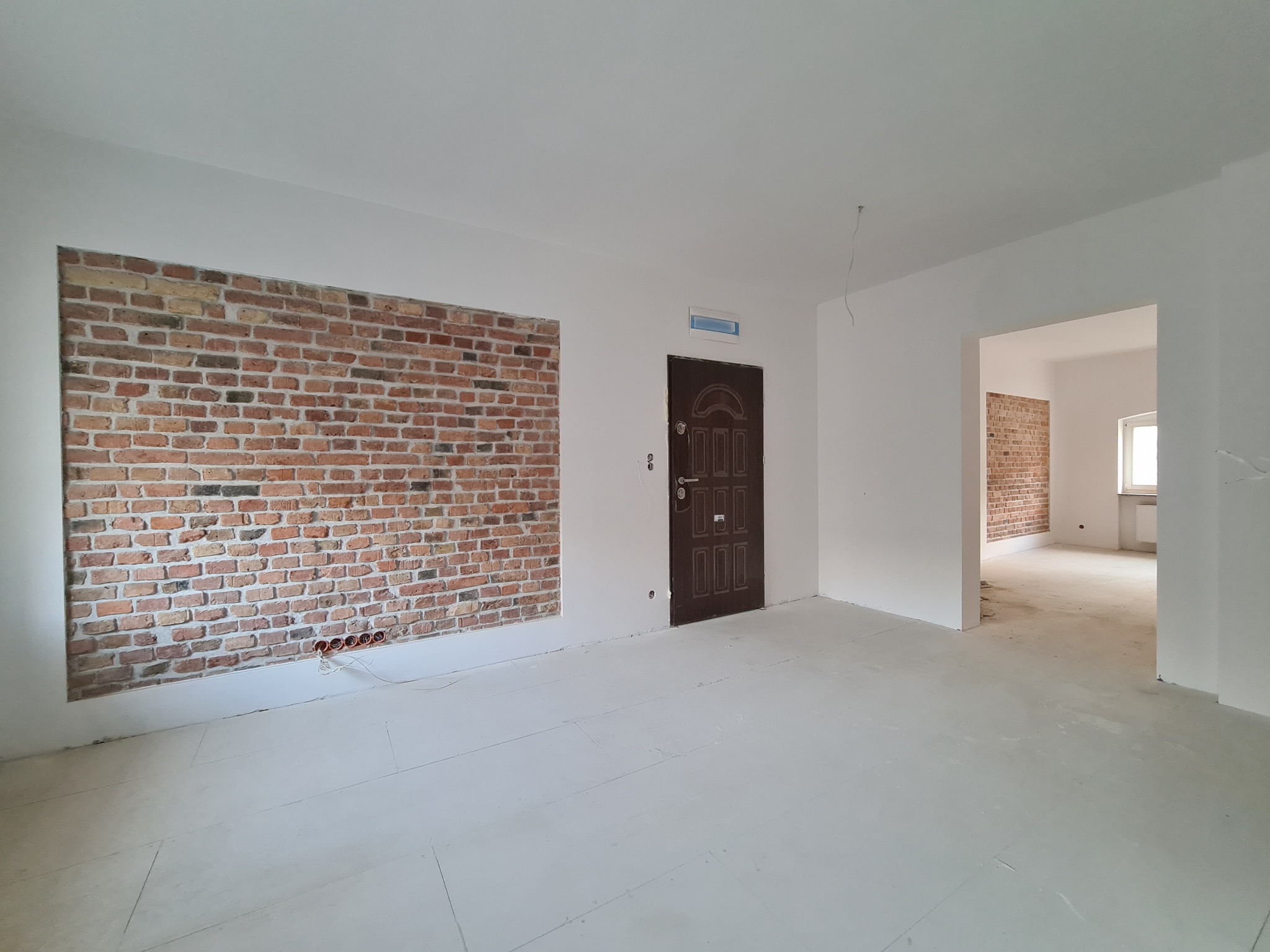 Wnętrze w trakcie remontu: fragment ściany z cegły dekoracyjnej, pomalowana podłoga, ciemne drzwi, widoczne przewody elektryczne.