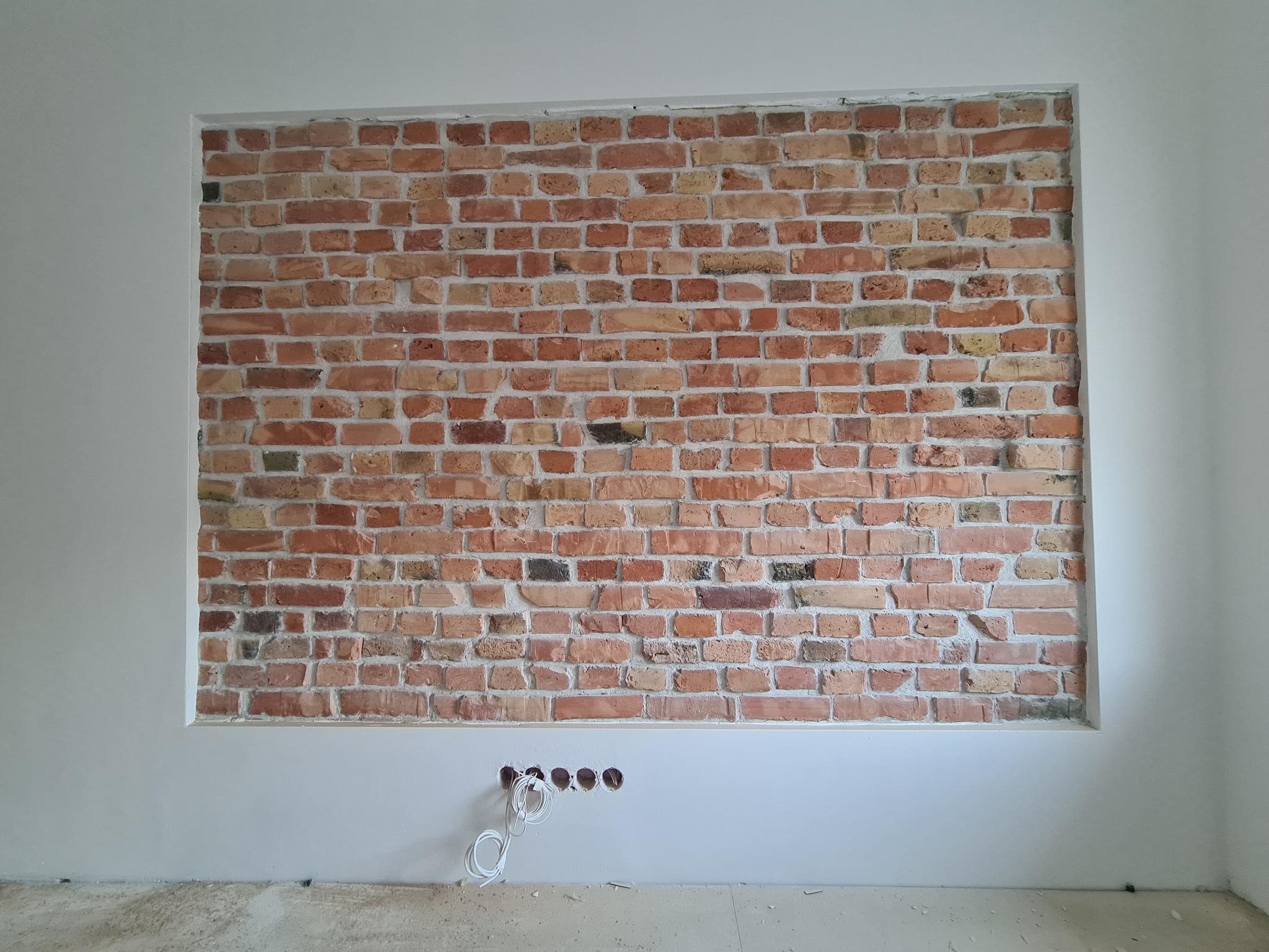 Fragment ściany z dekoracyjną cegłą, otoczony pomalowanym na biało fragmentem, w dolnej części widoczne otwory na kable.