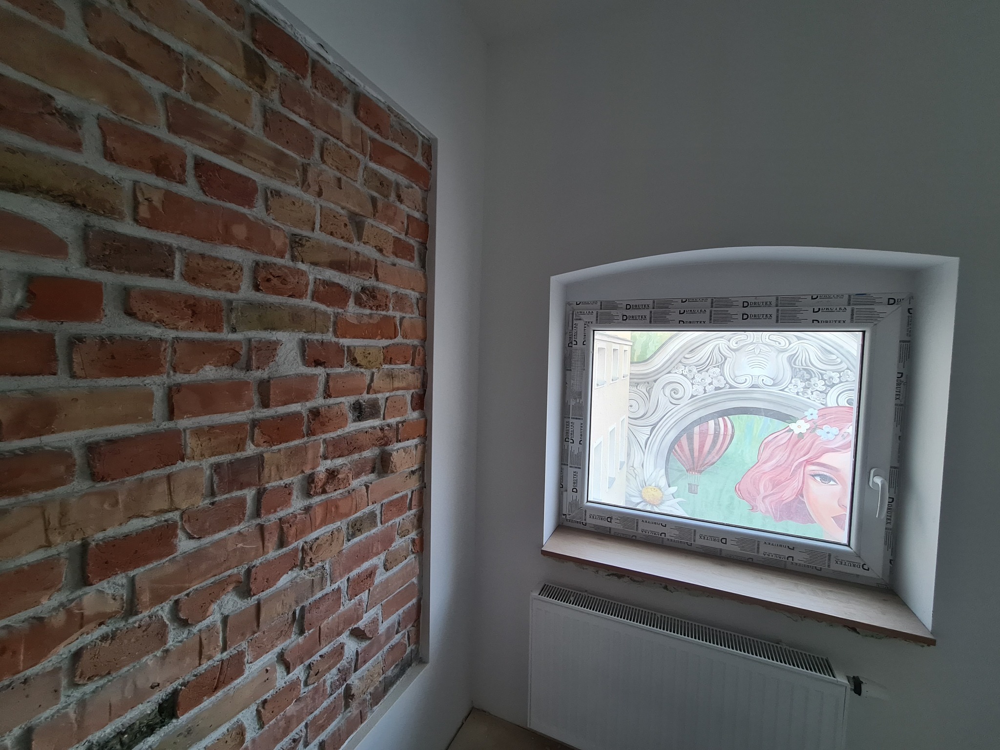Fragment wnętrza z odsłoniętą ceglaną ścianą i oknem z dekoracyjną folią przedstawiającą portret kobiety i balony