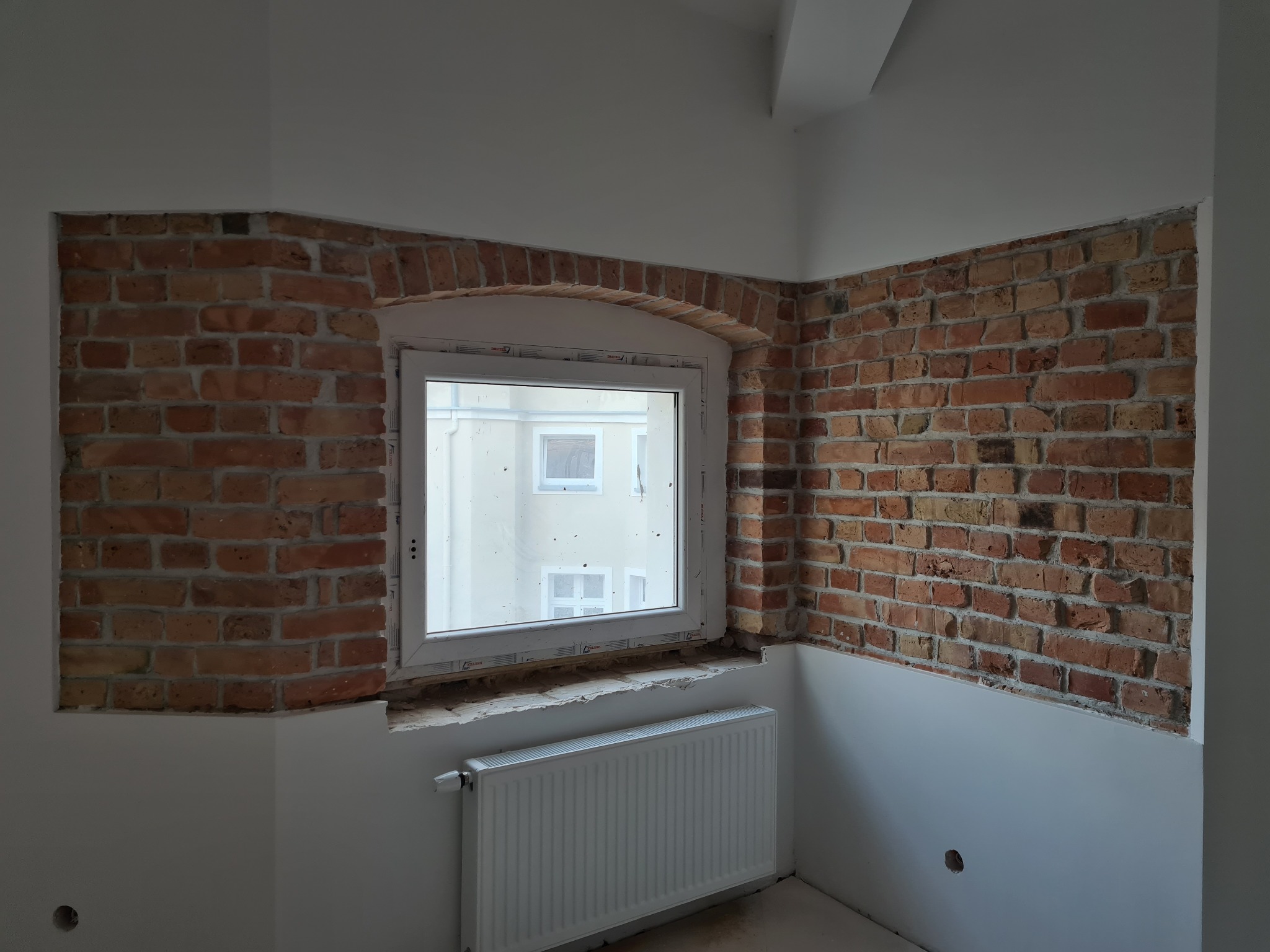 Fragment pokoju z oknem i odsłoniętą cegłą, grzejnik pod oknem, ściany częściowo otynkowane na biało.