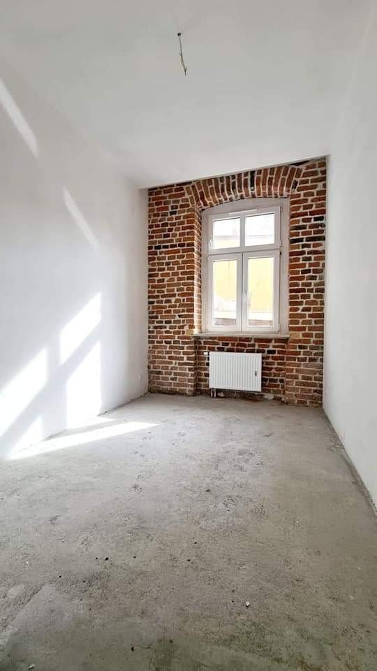 Wnętrze pokoju w trakcie remontu: surowe ściany, odsłonięta cegła wokół okna z białymi ramami, betonowa podłoga, biały grzejnik pod oknem.