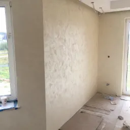 Ściana wewnętrzna z nałożoną dekoracyjną, beżową strukturą, zabezpieczona podłoga kartonem, widoczne puszki elektryczne i wystające przewody oświetleniowe.