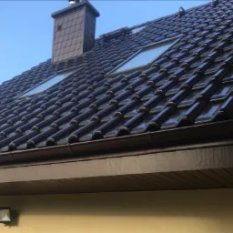 Błyszczący, brązowy dach z dachówki ceramicznej z dwoma oknami dachowymi i kominem, widoczny fragment elewacji budynku z oświetleniem zewnętrznym.