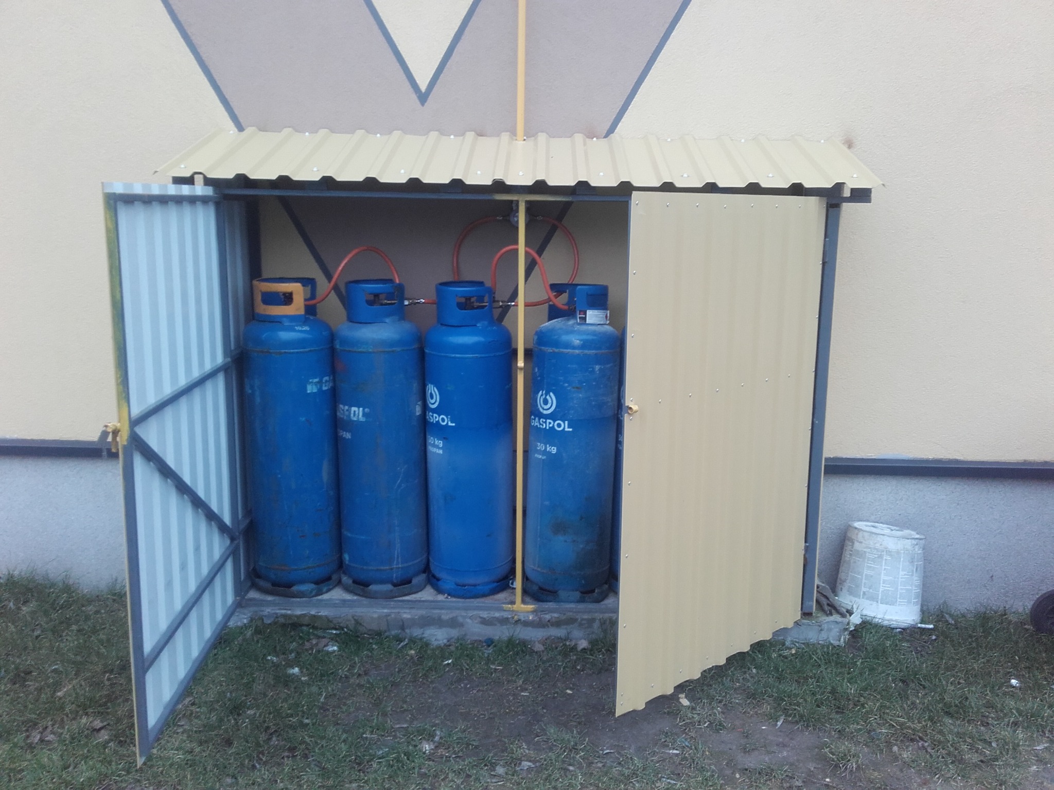 Zewnętrzna szafka z blachy falistej, mieszcząca cztery niebieskie butle gazowe GASPOL 30 kg, z otwartymi drzwiami z lewej strony i rurkami gazowymi u góry, na tle ściany budynku.