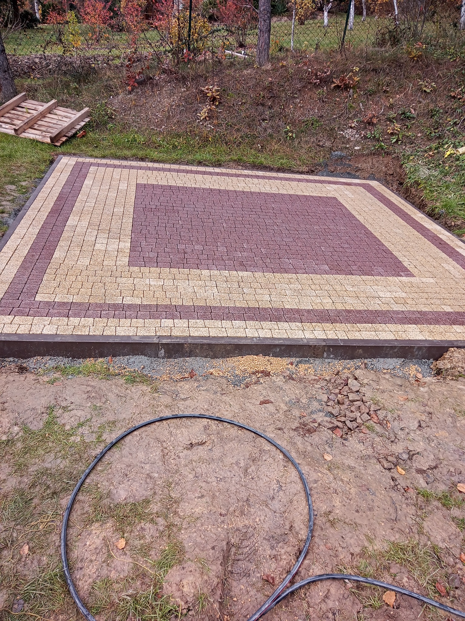 Nowo ułożony plac z kostki brukowej w dwóch kolorach, otoczony obrzeżem, widoczne przygotowane podłoże i fragment węża ogrodowego na błotnistym gruncie.