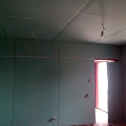 Marek Birula - Surowy pokój z płytami gipsowo-kartonowymi na ścianach i suficie, widoczne łączenia płyt, wisząca żarówka na kablu, przygotowany do dalszych prac wykończeniowych.