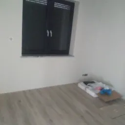 Ułożone panele podłogowe w jasnym odcieniu drewna w pokoju z czarnym oknem w Namysłowie, widoczne materiały budowlane w rogu pomieszczenia.