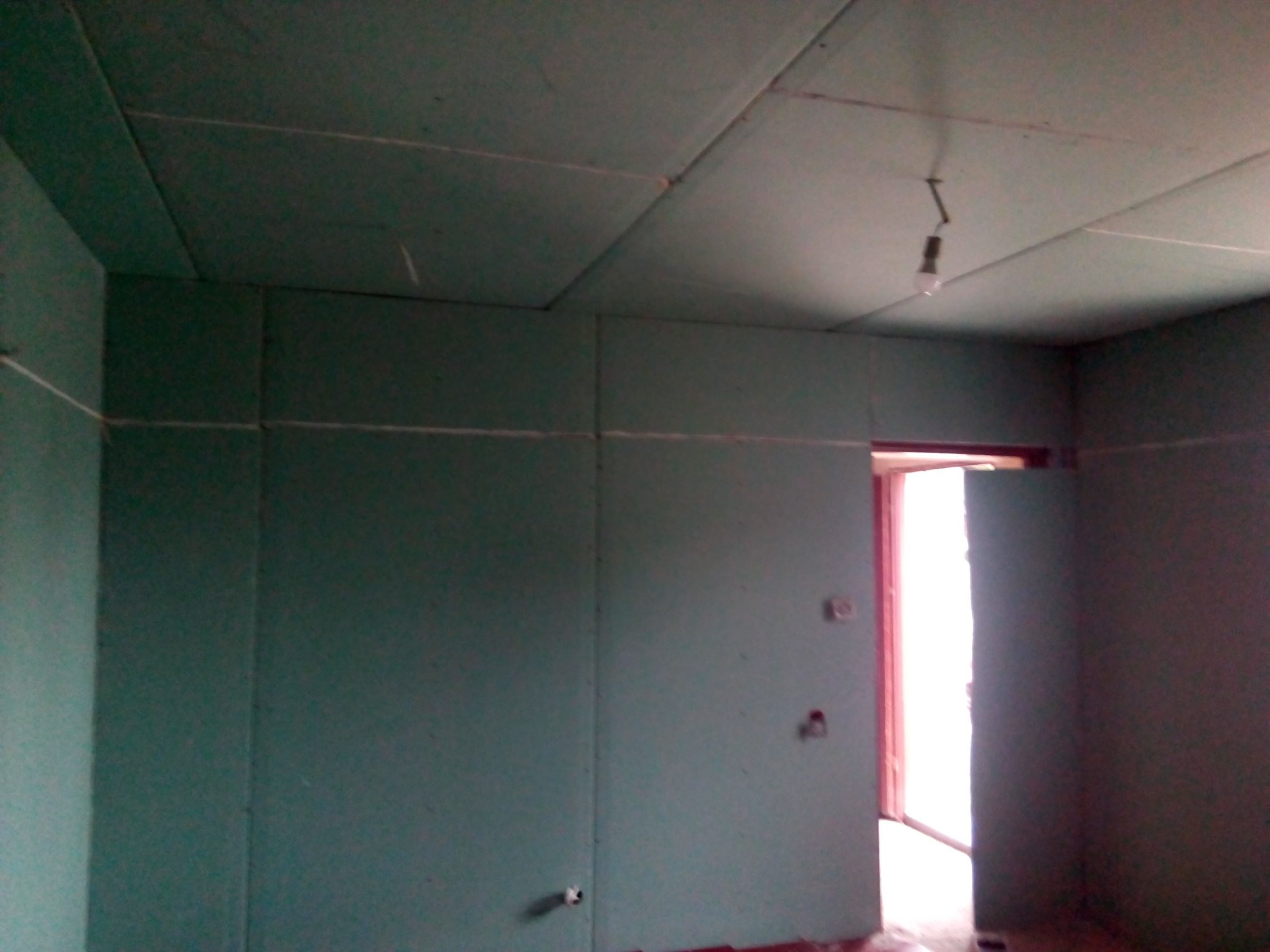 Surowy pokój z płytami gipsowo-kartonowymi na ścianach i suficie, widoczne łączenia płyt, wisząca żarówka na kablu, przygotowany do dalszych prac wykończeniowych.