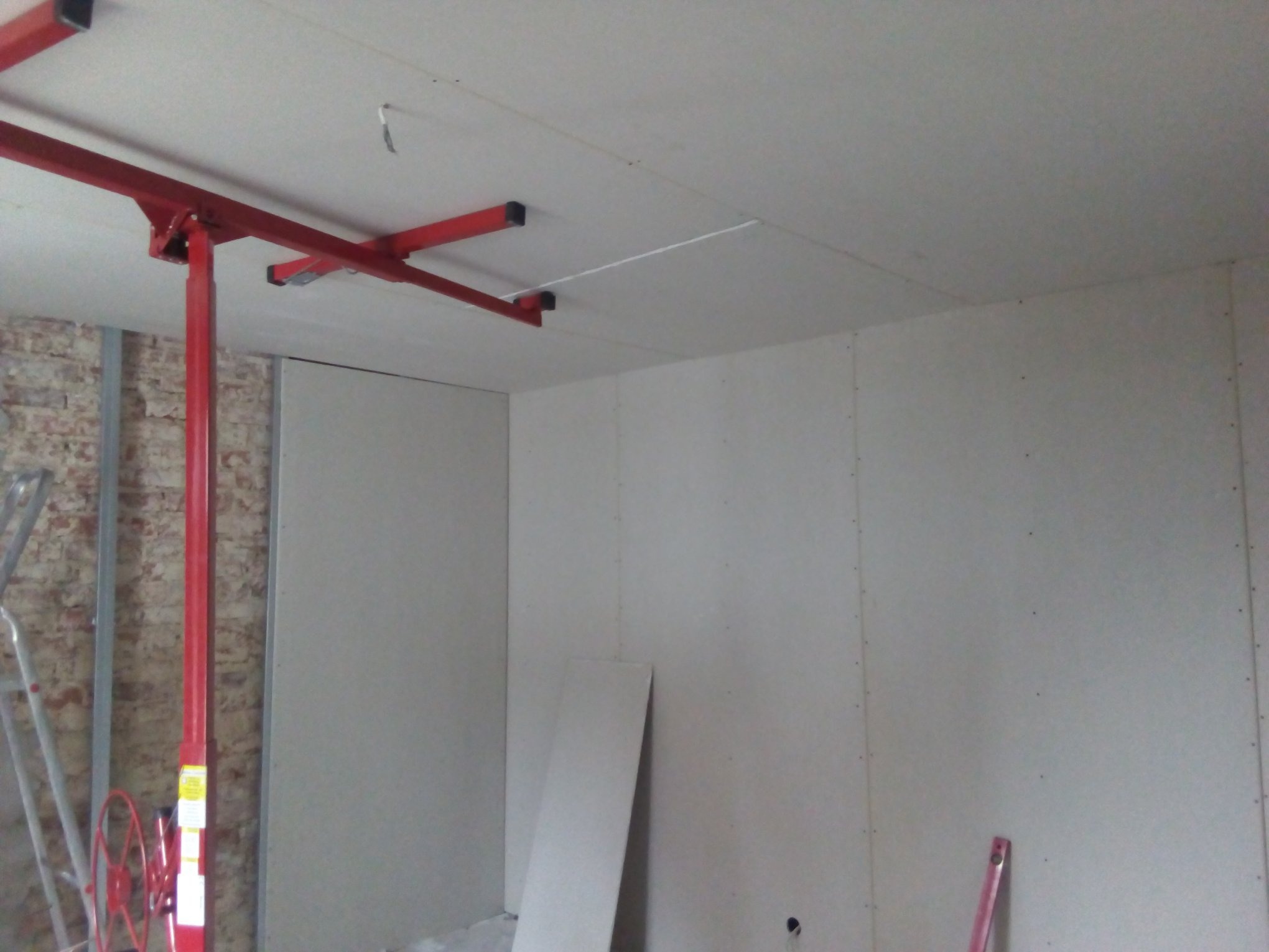 Montaż płyt gipsowo-kartonowych na suficie z użyciem podnośnika; widoczna surowa ceglana ściana w tle.