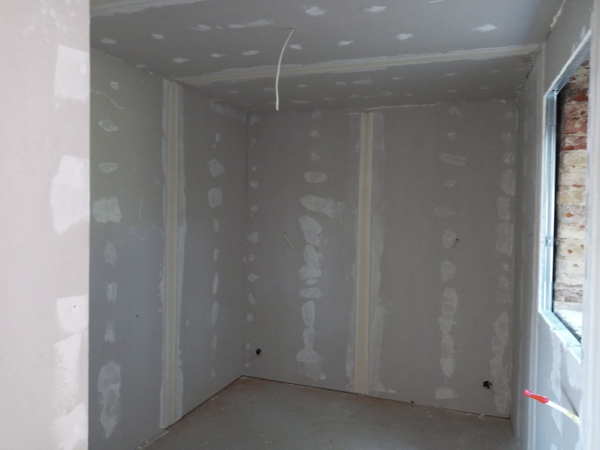 Wnętrze pomieszczenia w trakcie montażu płyt gipsowo-kartonowych, widoczne łączenia płyt pokryte masą szpachlową, wystający przewód elektryczny z sufitu, fragment okna z widoczną cegłą na zewnątrz.