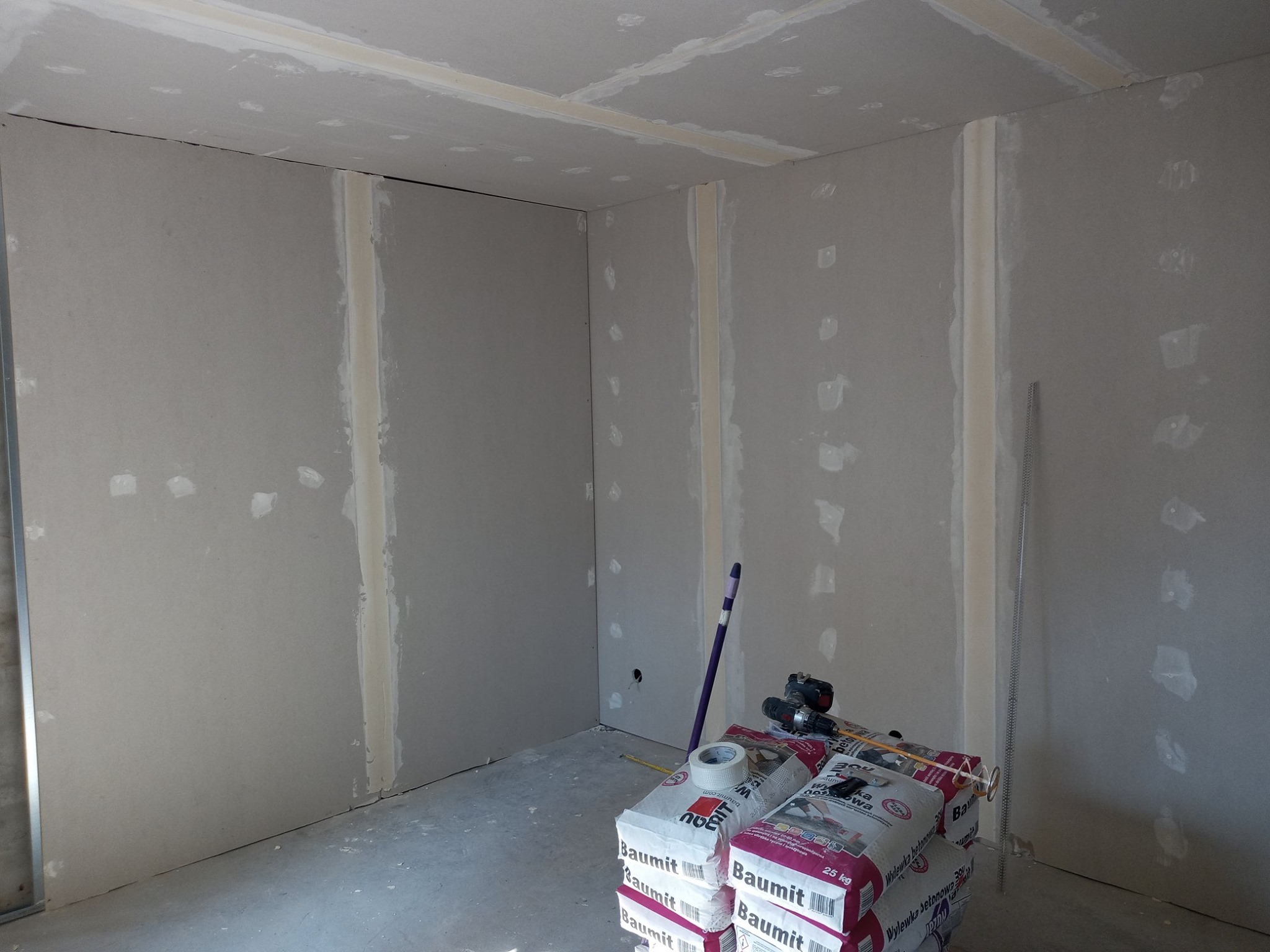 Wnętrze w trakcie prac wykończeniowych: ściany i sufit pokryte płytami gipsowo-kartonowymi, widoczne łączenia płyt wypełnione masą szpachlową, na podłodze worki z zaprawą, wiertarka i narzędzia.
