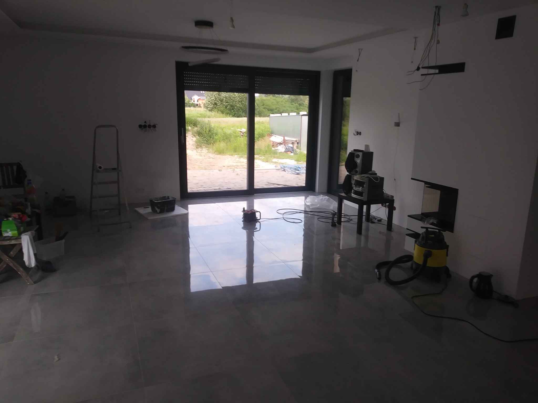 Wnętrze w trakcie remontu z dużym oknem tarasowym, widoczne narzędzia, drabina, podłoga z płytek oraz przygotowane ściany do malowania.