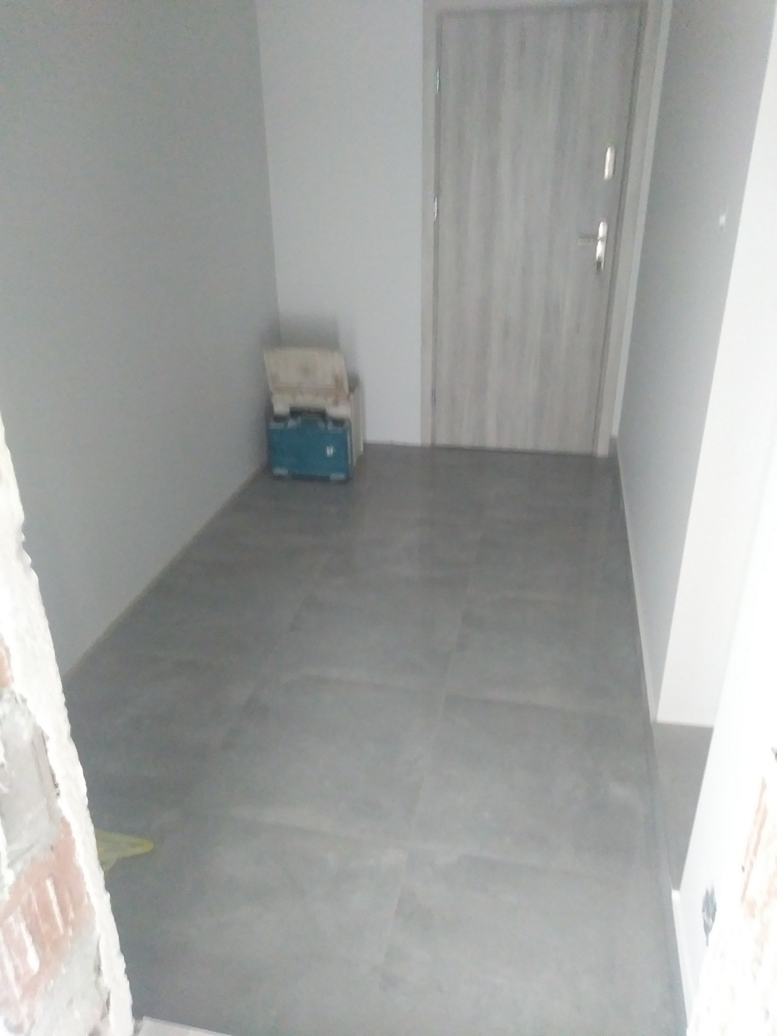 Szare płytki podłogowe ułożone w korytarzu, widoczna drzwi i skrzynka narzędziowa w rogu pomieszczenia, fragment surowej ściany z cegły w dolnym rogu kadru.