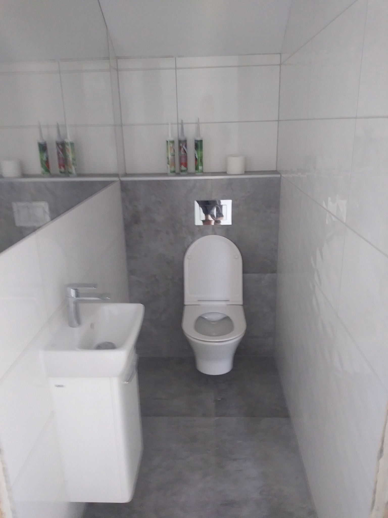 Nowoczesna łazienka z białą umywalką, toaletą i szarą ścianą akcentową. Widoczne tuby z silikonem na półce, sugerujące prace wykończeniowe.