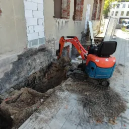 Pomarańczowo-niebieska mini koparka Kubota przy wykopie fundamentów budynku z odsłoniętą cegłą, zabezpieczonymi otworami okiennymi i świeżo ułożoną kostką brukową.