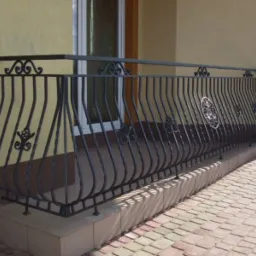 Czarna, metalowa barierka balkonowa z ozdobnymi elementami i rozetą na tle beżowej ściany budynku i kostki brukowej.