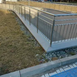 Zewnętrzna balustrada ze stali nierdzewnej zabezpieczająca zejście do piwnicy, widoczne elementy betonowe oraz otoczenie z kostki brukowej i trawy.