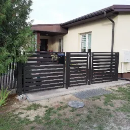 Nowoczesna, pozioma brama z metalowymi przęsłami w kolorze grafitowym, z czarną skrzynką na listy z numerem 79, na tle domu z beżową elewacją i brązowymi elementami wykończeniowymi, widoczny...