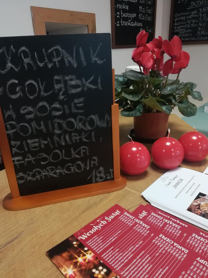 Tablica kredowa z wypisanym menu: krupnik, gołąbki w sosie pomidorowym, ziemniaki, fasolka szparagowa, cena 18 zł, obok czerwone świece, ulotki świąteczne i menu restauracji na drewnianym stole