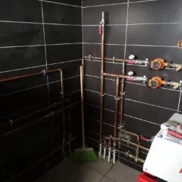 Miedziane rury instalacji grzewczej z manometrami i pompami obiegowymi na tle czarnych płytek, widoczny również piec grzewczy Heitz z 3-letnią gwarancją oraz akcesoria instalacyjne i miotła.