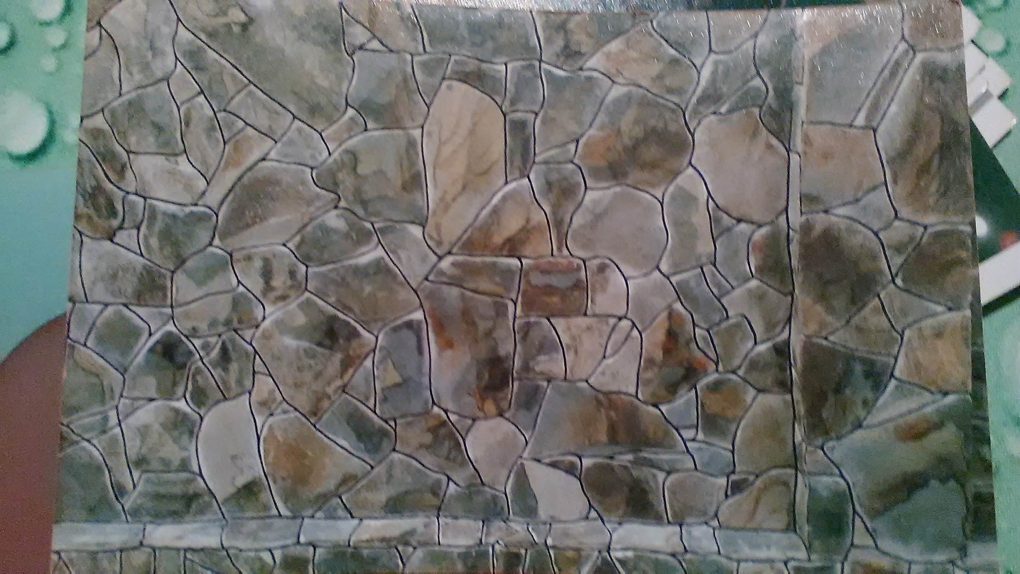 Ściana wyłożona płytkami imitującymi kamień w odcieniach szarości i brązu, z wyraźnymi fugami. Płytki ułożone nieregularnie, tworząc efekt naturalnego kamiennego muru.