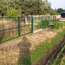 Zielone panele ogrodzeniowe z siatki zamontowane na betonowej podmurówce, widok z zewnątrz posesji, w tle samochód z przyczepką i osoba.