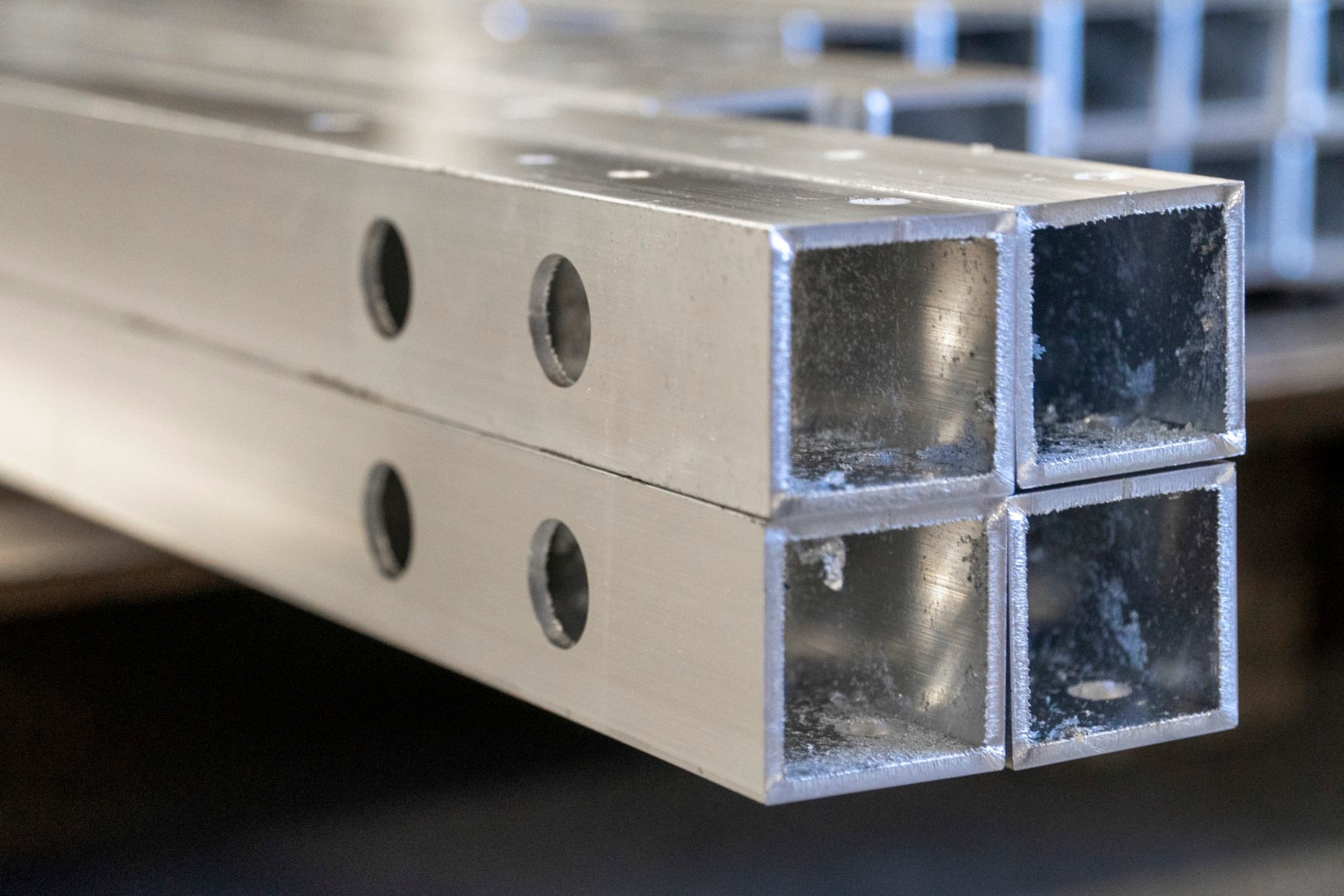 Dwa kwadratowe, metalowe profile z nawierconymi otworami, ułożone jeden na drugim, widoczne ślady obróbki metalu wewnątrz profili.