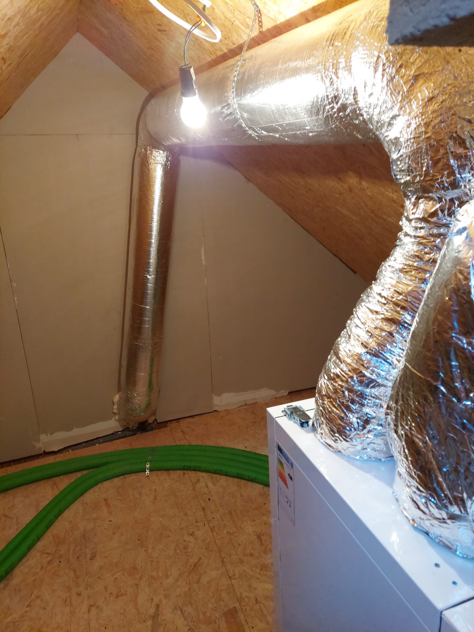 Instalacja systemu wentylacji z rekuperacją na poddaszu: widoczne srebrne rury izolowane termicznie, zielone przewody wentylacyjne i centrala rekuperacyjna z etykietą energetyczną, oświetlone żarówką.