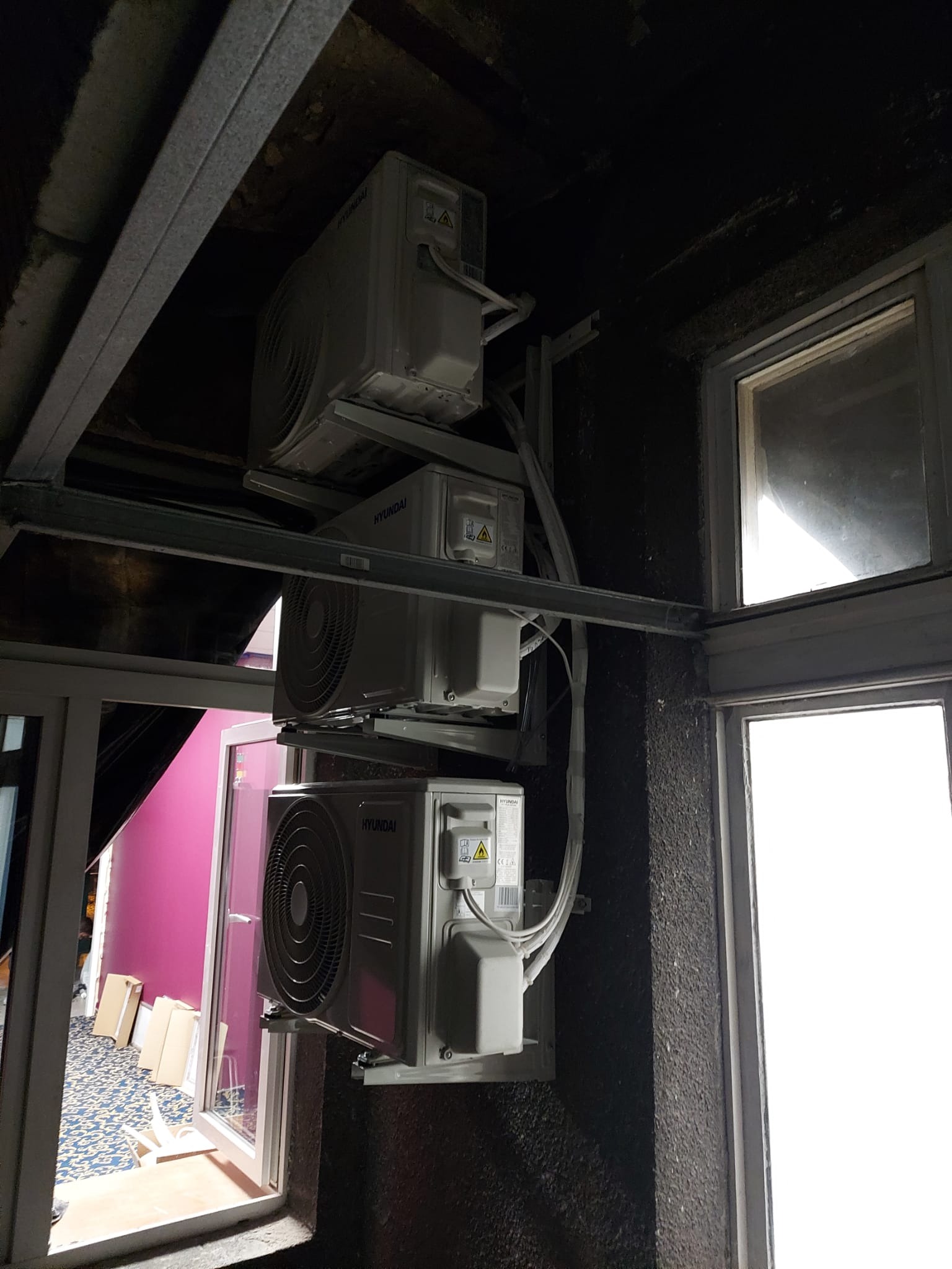 Trzy jednostki zewnętrzne klimatyzacji Hyundai zamontowane jedna nad drugą na metalowej konstrukcji przy oknie, widok z dołu na ciemne, surowe wykończenie sufitu i ściany.