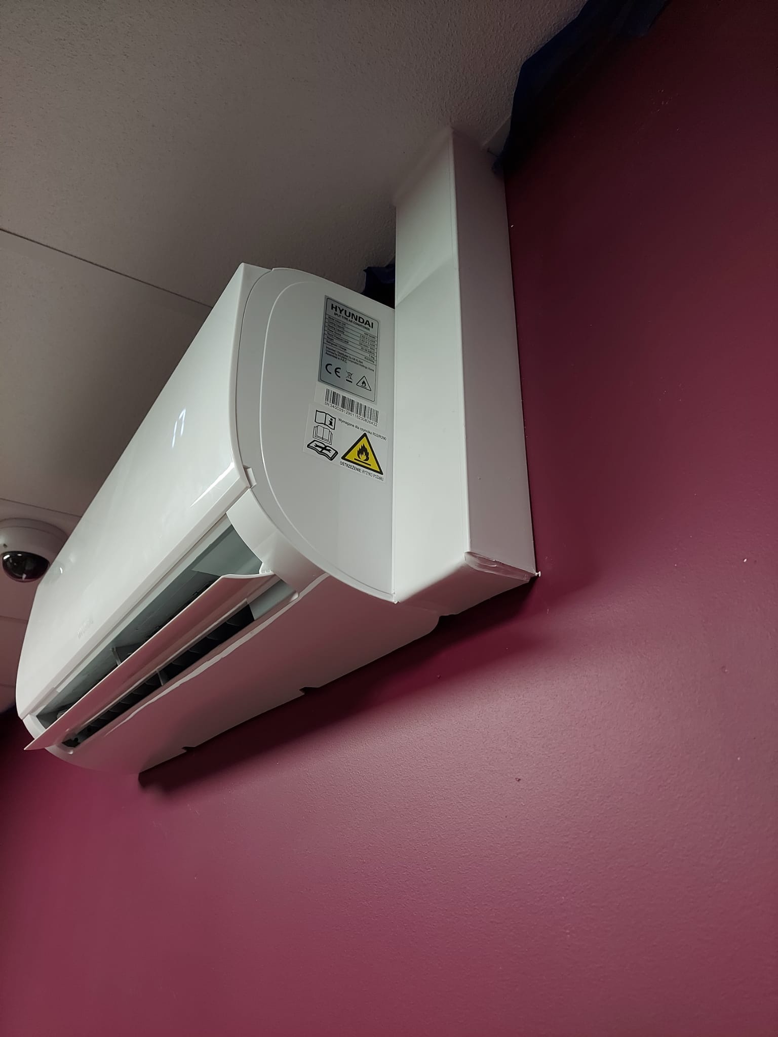 Biały klimatyzator marki Hyundai zamontowany na bordowej ścianie, widoczna naklejka ostrzegawcza i wyświetlacz temperatury ustawionej na 17 stopni, w tle widoczny fragment kamery monitoringu.