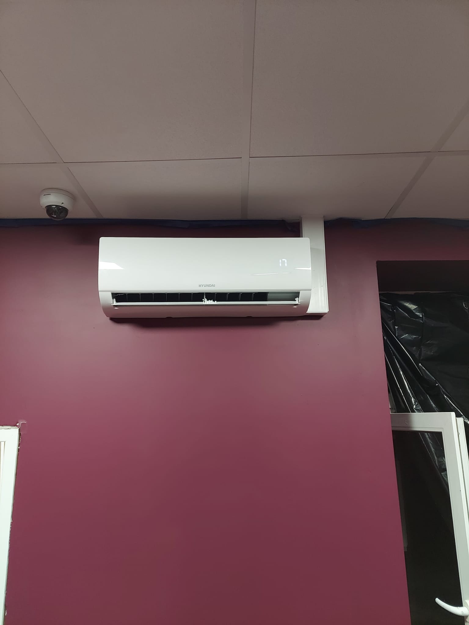 Biały klimatyzator Hyundai zamontowany na bordowej ścianie w pomieszczeniu z sufitem kasetonowym i kamerą monitoringu.