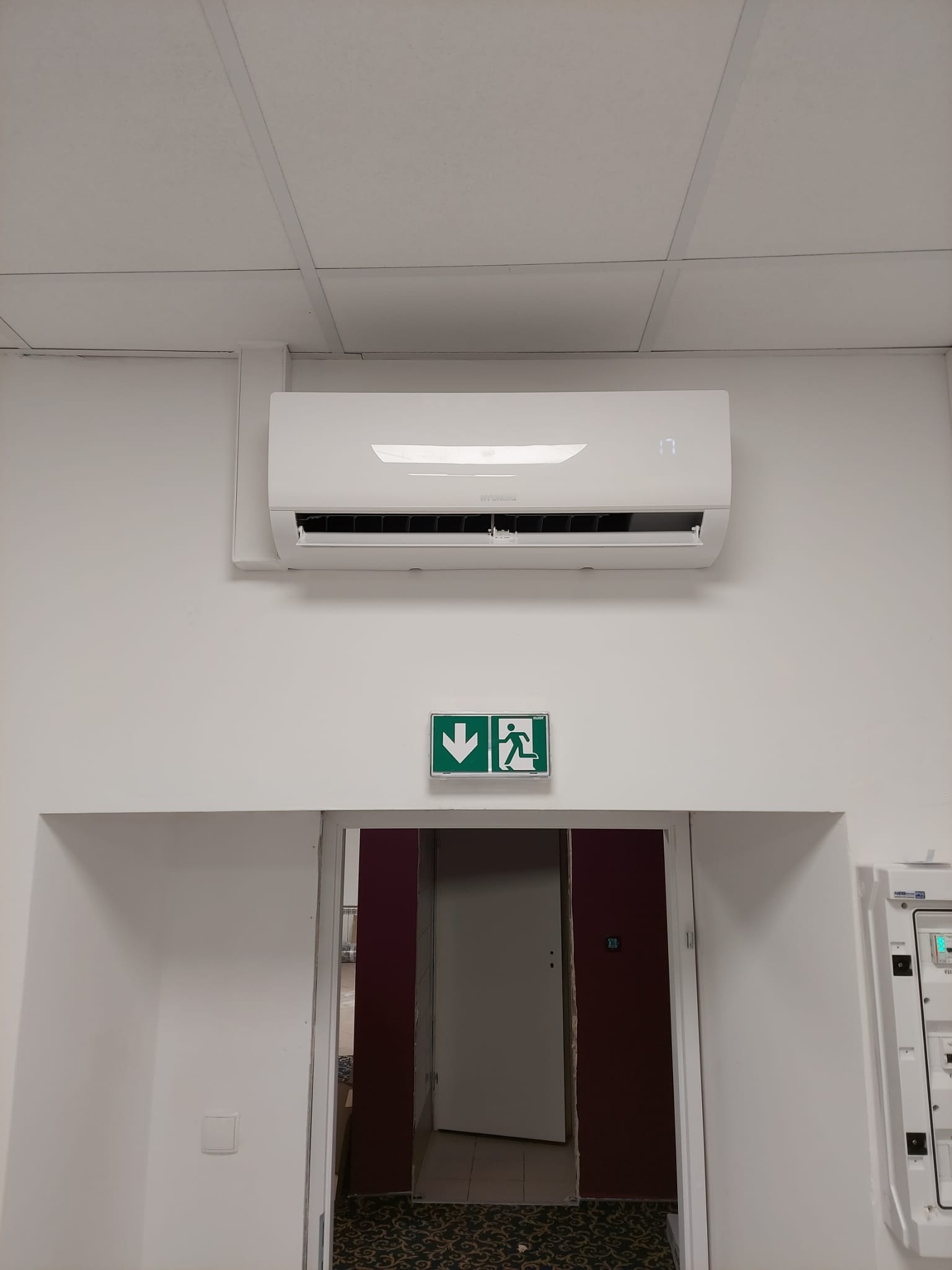 Biały klimatyzator ścienny z wyświetlaczem temperatury (17 stopni Celsjusza) zamontowany nad drzwiami ewakuacyjnymi oznaczonymi zielonym znakiem z piktogramem biegnącej osoby.