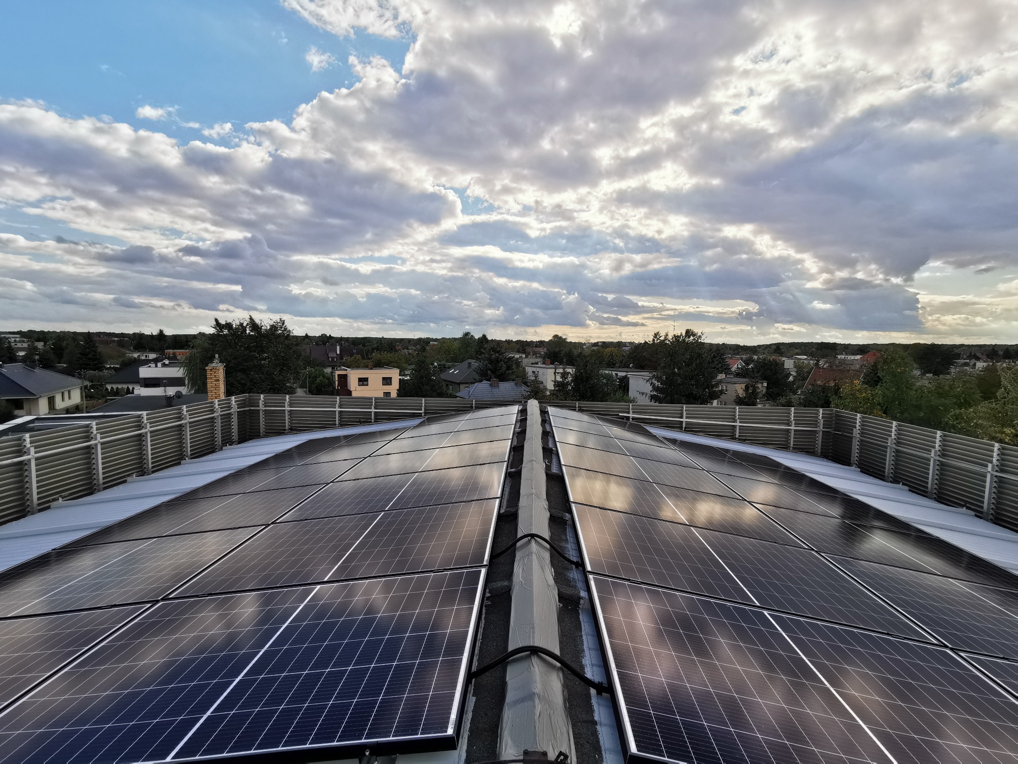Widok z góry na instalację paneli słonecznych na dachu budynku mieszkalnego, z widocznym krajobrazem miejskim i zachmurzonym niebem.