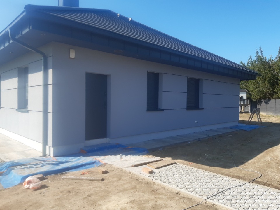 Nowoczesny dom jednorodzinny z szarą elewacją, czarnym dachem i oknami z roletami zewnętrznymi, widoczne zabezpieczenia folią malarską przy elewacji i chodniku z ażurowych płyt.