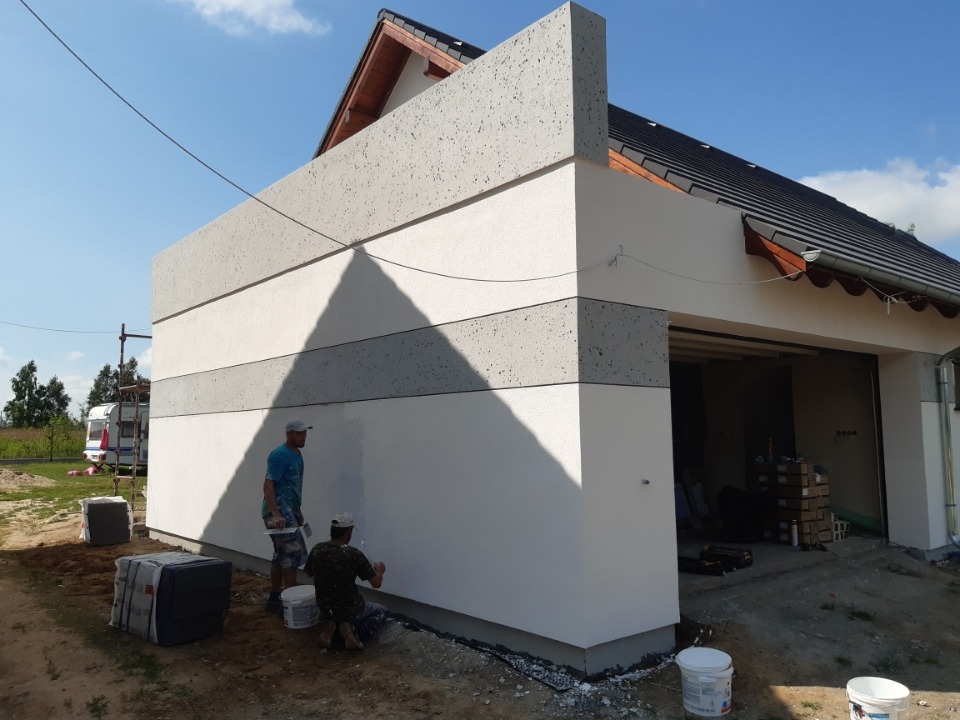 Dwóch mężczyzn tynkuje elewację budynku z garażem, widoczne pasy tynku w odcieniach szarości i bieli, niezadaszony teren budowy.